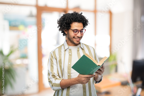 knowledge, education and people concept - happy smiling young man in glasses reading book over office background