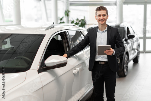 Cheerful young sales manager with tablet computer standing near new automobile at luxury car salon, copy space