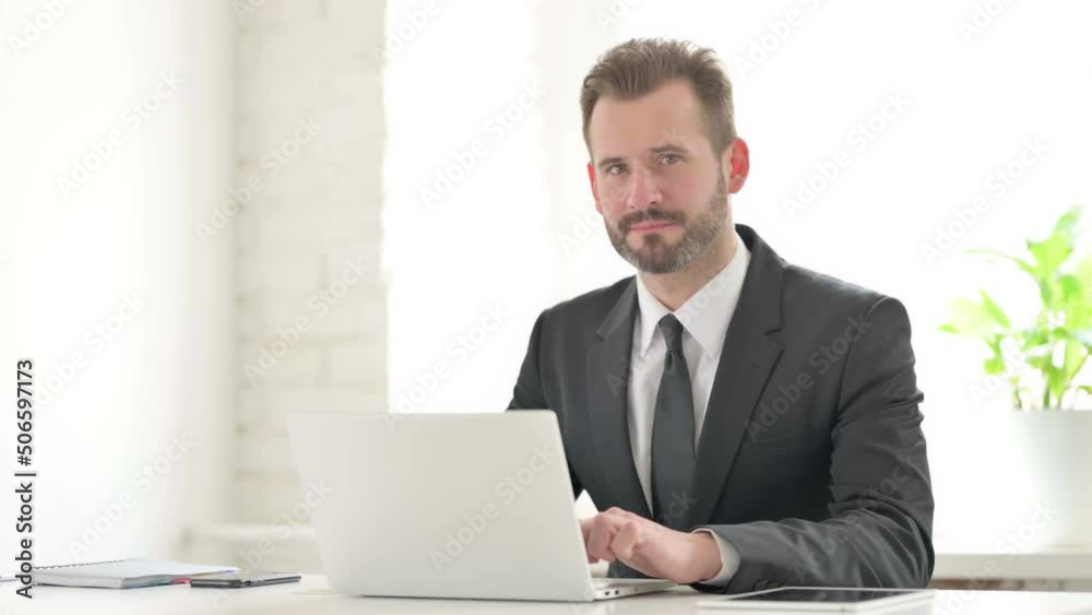 Young Businessman Smiling at Camera while using Laptop in Office Young Businessman Smiling at Camera while using Laptop in Office