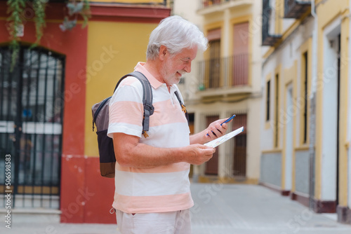 Happy senior tourist man holding backpack on shoulder walking in the alleys of the old town in Seville, Spain while following map direction on mobile phone photo