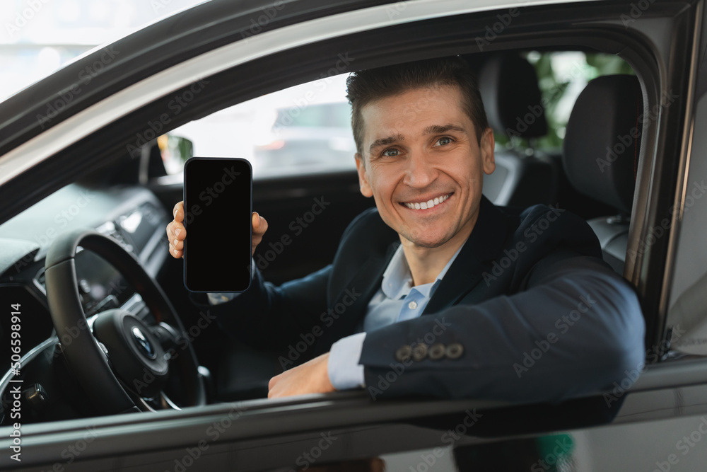 Handsome young car salesman sitting in auto salon, showing blank mobile phone with mockup for design at dealership