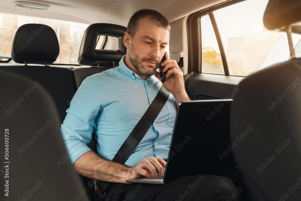 Businessman using laptop talking on smartphone while going by car