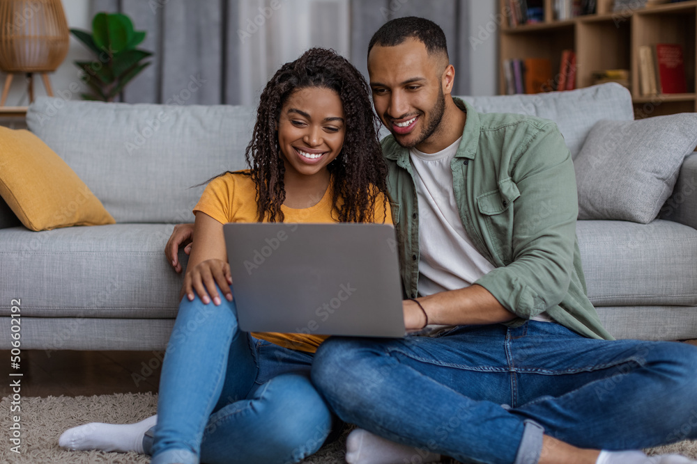 Positive african american lovers sitting on floor by couch with laptop, websurfing and having fun together at home