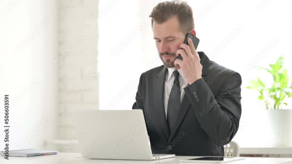 Young Businessman Talking on Phone while using Laptop in Office Young Businessman Talking on Phone while using Laptop in Office