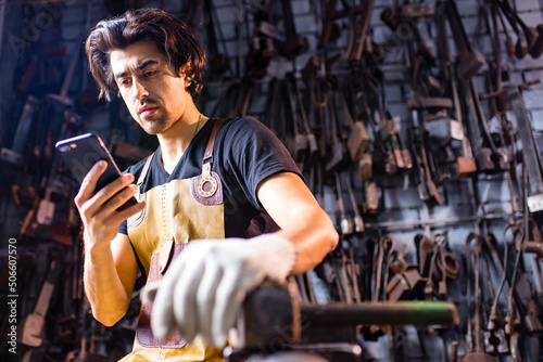 Authentic blacksmith man wear leather apron in dark indoors studio looking at phone screen