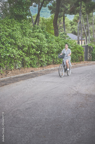 Young adult woman with a bow in her hair and a striped dress enjoys a beautiful summer day riding a bike. She pedals down a quiet street.
