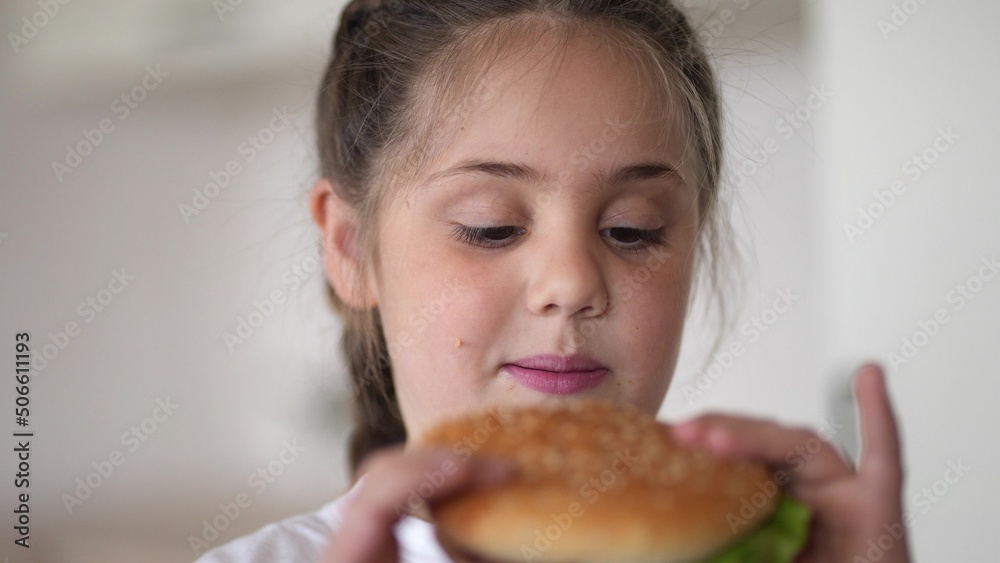 little girl eating a hamburger. unhealthy fast food proper lifestyle nutrition concept. child greedily with pleasure bites a big burger in the kitchen at home. kid eats fast food close-up