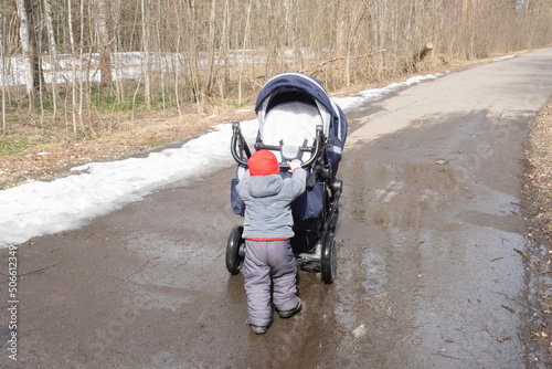 Small child in red cap pushing double blue pram on wet asphalt on the background of spring forest