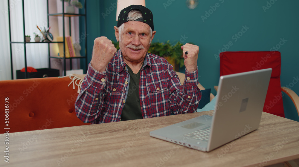Φωτογραφία Overjoyed senior grandfather man on home sofa with laptop scream in delight raise hands in triumph winner gesture celebrate success win money in lottery