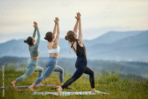 Back view of young females in sportswear rising hands up joining together practicing virabhadrasana in morning mountains. Beautiful women standing in warrior pose outdoors. Concept of health. photo