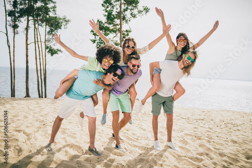Full length photo of crazy positive friends piggyback raise hands up enjoy warm weather sand beach outdoors