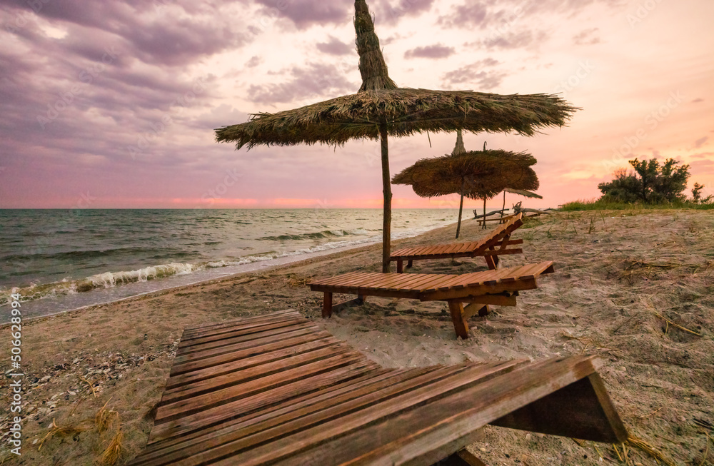 Beautiful sunrise or sunset over the calm waters of the Black Sea on the Kinburg Spit in Ukraine. Beach summer landscape. Straw umbrella and wooden deck chair on the beach at sunset or sunrise