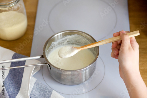woman's hand holds a wooden spoon of porridge over a pot. Cooking semolina on a white electric stove