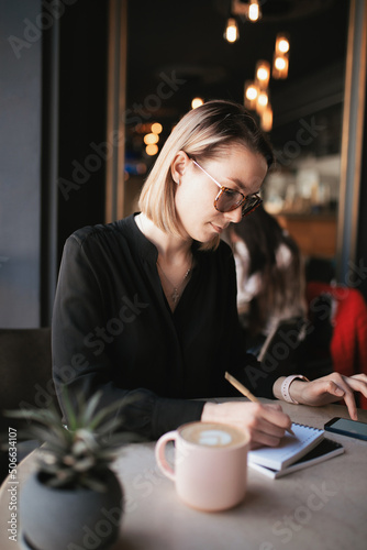 A woman works remotely, looking for information on her smartphone and making notes in a notebook.