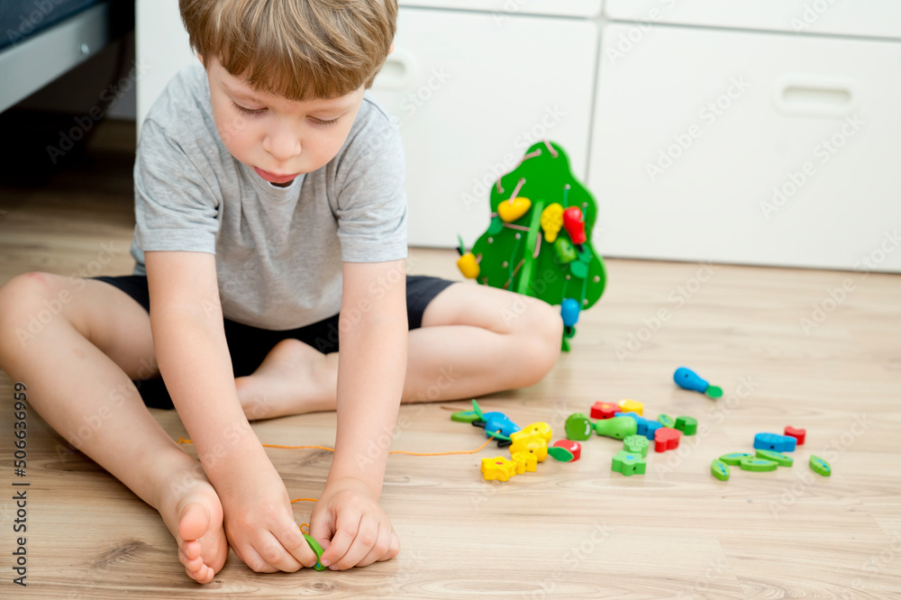 Fine-motoric skils. Stringing Beads Toys for infants and toddlers. Threading Activity for Children. boy sitting on the floor and plays with wooden tree and garden goods. Fine-motoric skils. Stringing Beads Toys for infants and toddlers. Threading Activity for Children. boy sitting on the floor and plays with wooden tree and garden goods.