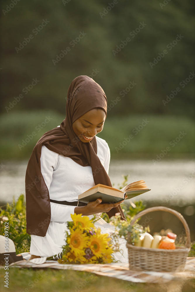© prostooleh - A muslim woman sits on the plaid picnic blanket near the river and reading a book © prostooleh - A muslim woman sits on the plaid picnic blanket near the river and reading a book