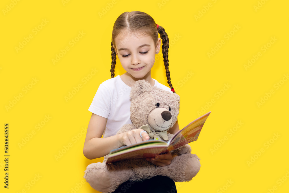 Girl child reads a book and holds a teddy bear on a yellow background ...