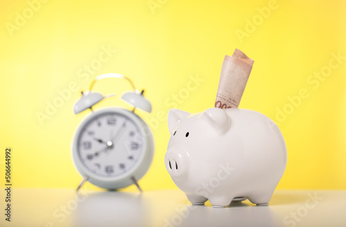 Banknotes are wearing white piggy banks and alarm clock on a table with yellow background, business, investment, finance, and Money Saving for the future concepts, time and money savings.