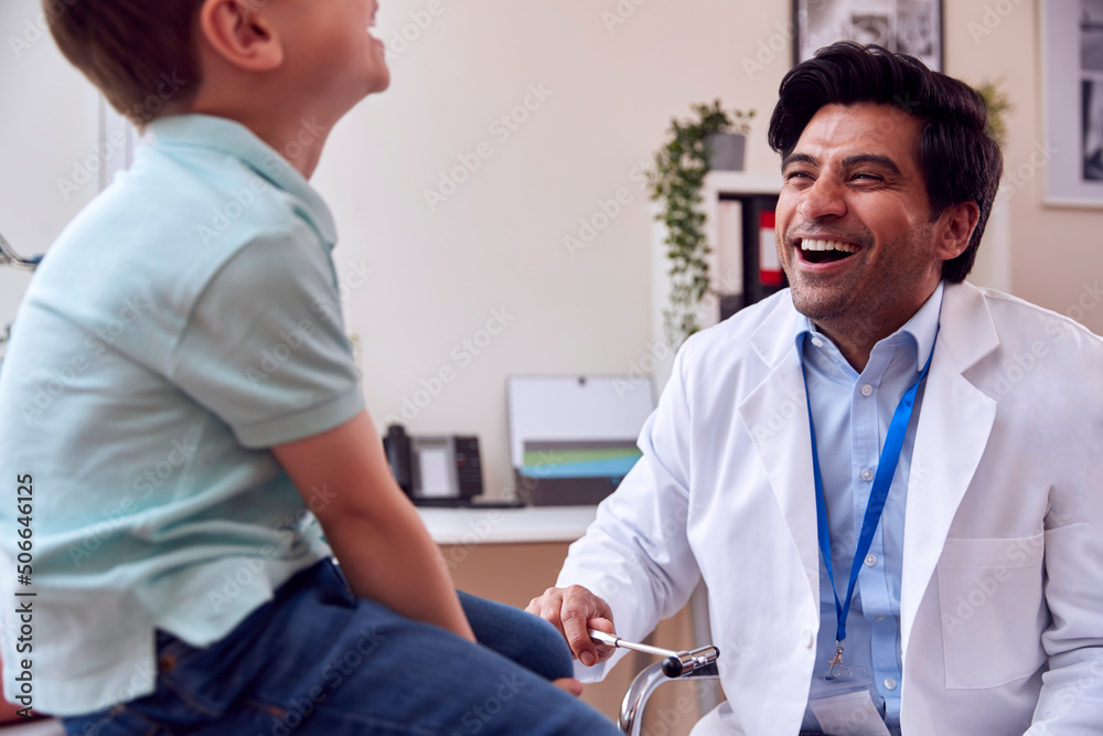 Fototapeta premium Male Doctor Or GP Wearing White Coat Examining Smiling Boy Testing Reflexes