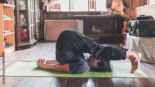 young Asian man practicing yoga in a wooden room sitting in pose Plow Pose on a green yoga mat at a wooden house. healthy life concept