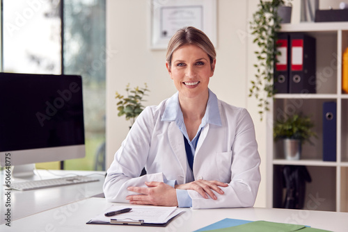 Portrait Of Smiling Female Doctor Or GP Wearing White Coat In Office Sitting At Desk