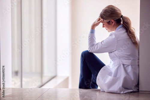 Stressed Female Doctor Wearing White Coat Sitting On Floor In Hospital Corridor With Head In Hands