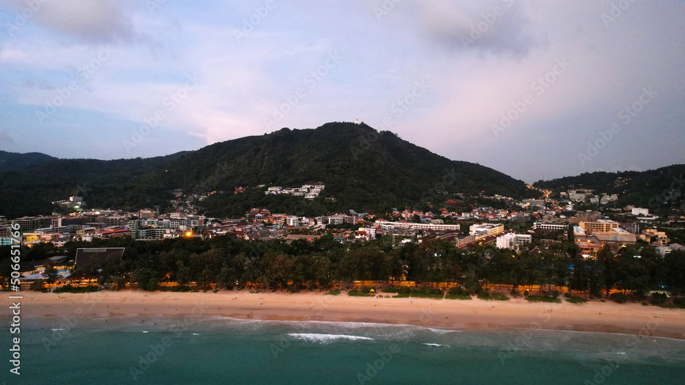 View of the beach and the island from a height. Sunset. People walk on the beach, relax. There are clouds in the sky. Green hills on the island of Phuket. There are hotels, lanterns are lit. Journey