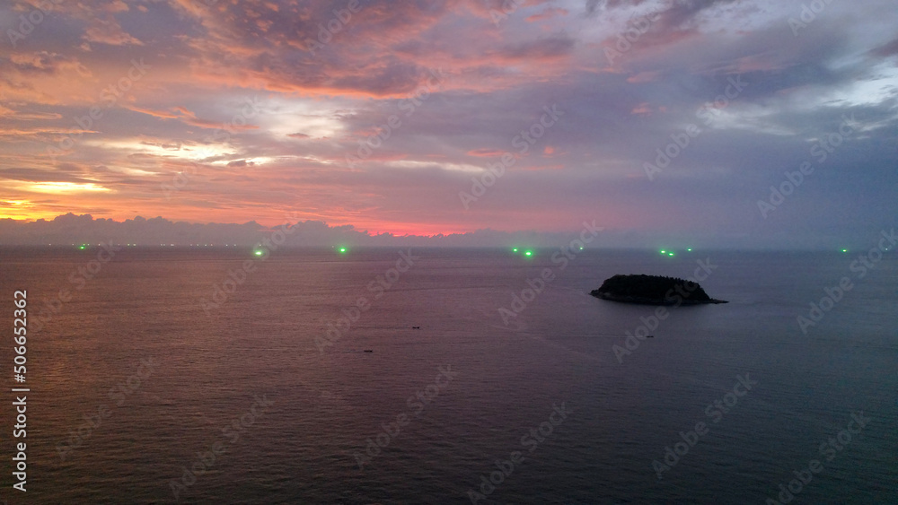 The red-orange sunset is reflected with clouds in the sea. An island can be seen in the distance. Smooth water is like a mirror. There is a lonely island. The green lights of fishing boats are shining