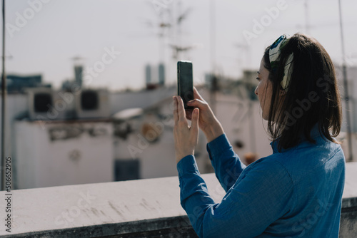 Woman with blue shirt and hairband taking photos with her phone on the roof of a building in Barcelona. Blurred building rooftops in the background. Film effect. Landscape.