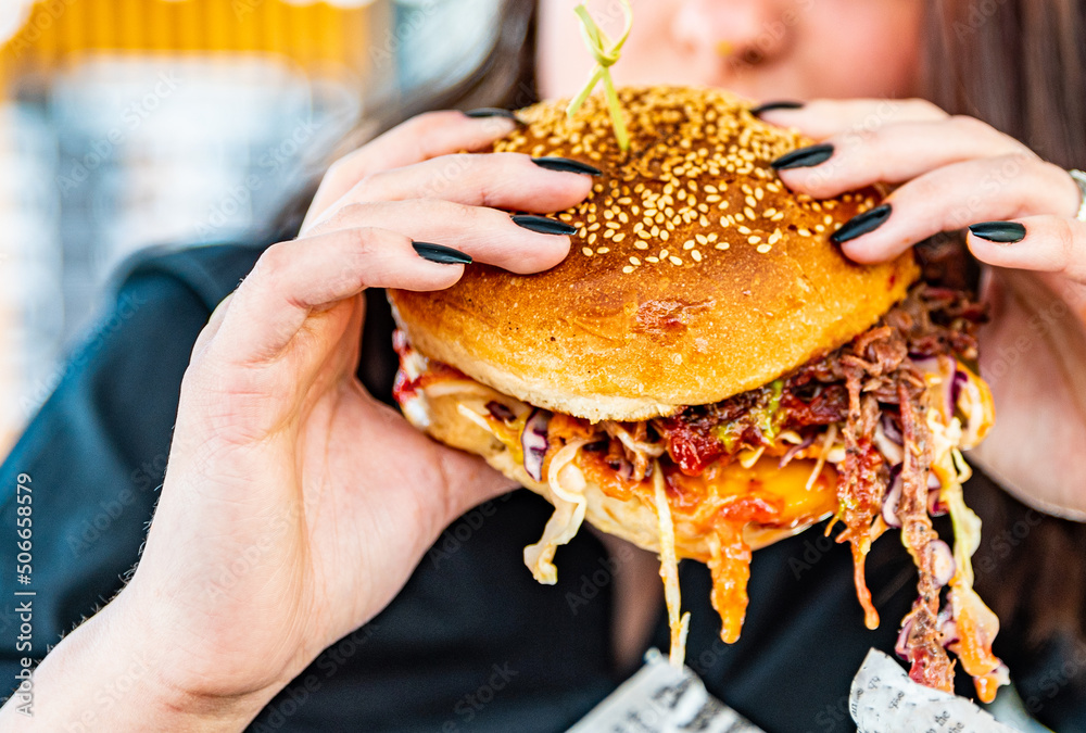 Woman eating street food burger outdoors. Traditional barbecue pulled beef burger with vegetables in woman hand Woman eating street food burger outdoors. Traditional barbecue pulled beef burger with vegetables in woman hand