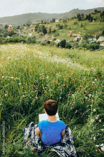 The boy sits in the grass on the mountain and draws a picture on the top of the mountain.