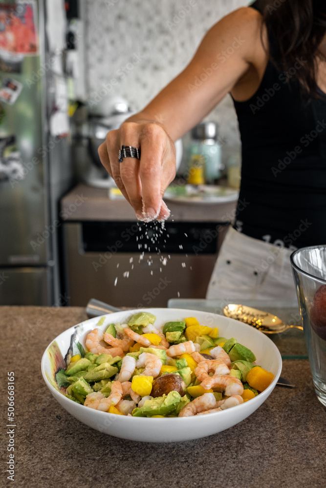Female hand adding salt to avocado and shrimp salad in a bowl. Seasoning ready meal by sprinkling salt. Woman preparing food at home kitchen. Organic healthy dieting cooking and losing weight concept.