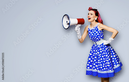 Purple haired woman holding red megaphone, shout advertising something. Girl in blue pin up style with mega phone loudspeaker. Grey color background with mock up. Female model in retro fashion dress.