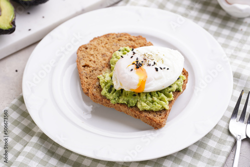Slices of gluten-free sunflower seeds bread with mashed avocado, poached egg and sesame seeds on white plate on green checkered napkin