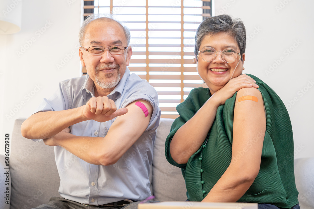 Obraz premium Coronavirus Vaccination, happy asian elderly, aged family smile strong together, showing bandage on arm with protect of covid-19 after injection of vaccine, sitting on couch in living room at home.