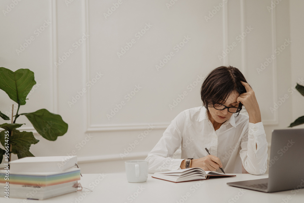 Fair-skinned adult woman sits at desk thoughtfully writing down important information in notebook. Brunette with glasses uses laptop. Working atmosphere concept