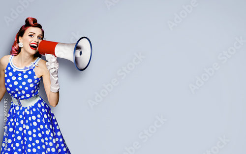 Portrait of purple haired woman holding mega phone, shout advertising something. Girl in blue pin up style dress with mega phone loudspeaker. Isolated on grey background. Beauty model in retro concept