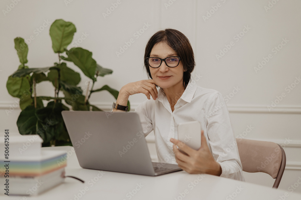 Successful adult caucasian brunette woman using notebook, work from home. Lady in glasses and shirt holds phone in her hands. Lifestyle concept, freelancing Successful adult caucasian brunette woman using notebook, work from home. Lady in glasses and shirt holds phone in her hands. Lifestyle concept, freelancing