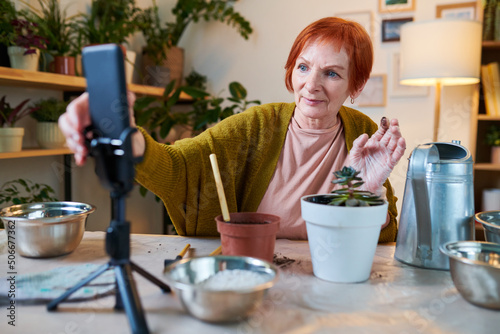 Mature woman sitting at table with potted flowers, bowls and watering can and talking to her followers online during stream on smartphone