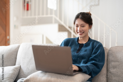 Happy casual beautiful young asian woman working on a laptop sitting on the couch in the house.