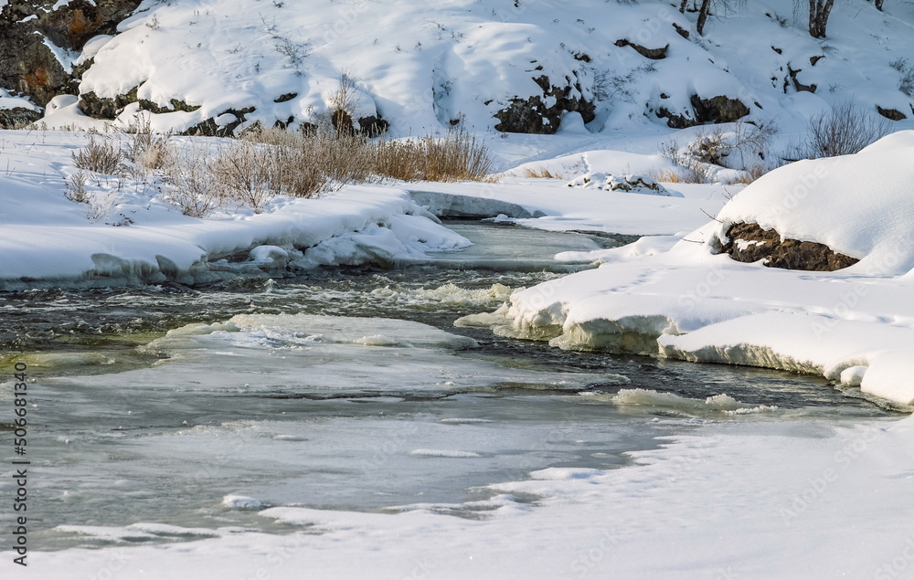 Winter landscape with a fast river with ice-free water, snow, dry grass and rocky shores