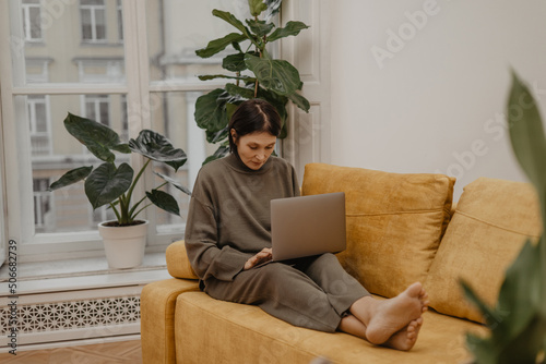 Photos Attentive caucasian adult woman works at home using notebook while sitting on comfortable sofa