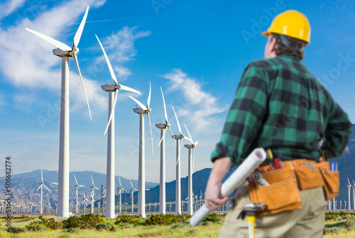 Male Contractor Wearing Tool Belt and Hard Hat Facing Alternative Energy Wind Turbines.