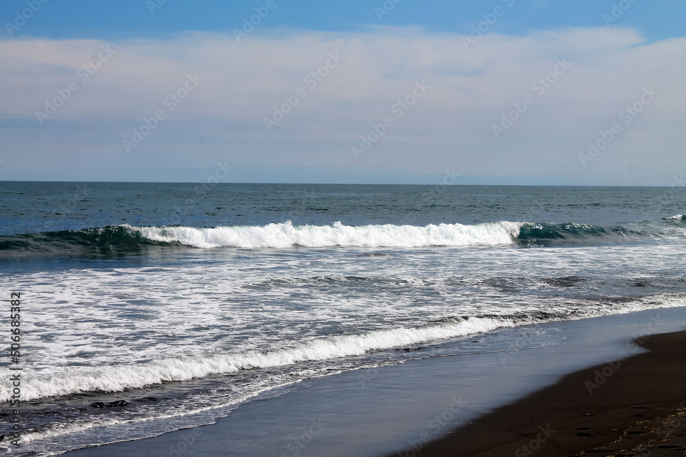 The coast of the Pacific Ocean is covered with black volcanic sand. Long turquoise surf waves roll and foam. Blue sky. Kamchatka. Khalaktyrsky beach. High quality photo