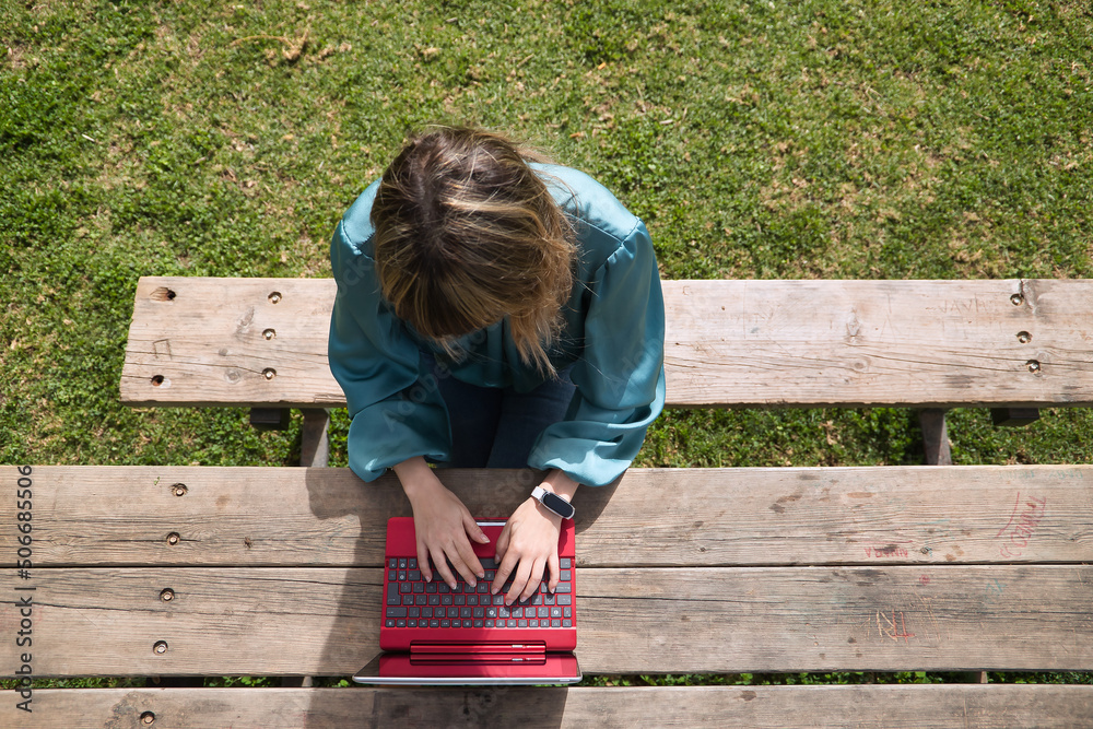 Young, pretty, blonde woman in green shirt, sitting on a wooden bench in a park, working with her laptop, view from above. Technology concept, tablet, notebook, teleworking, telecommuting, zenithal. Young, pretty, blonde woman in green shirt, sitting on a wooden bench in a park, working with her laptop, view from above. Technology concept, tablet, notebook, teleworking, telecommuting, zenithal.