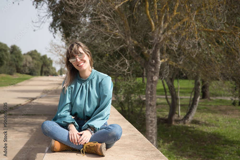 Fototapeta premium Young, pretty, blonde woman in green shirt, jeans and mirrored sunglasses, sitting smiling in a park. Concept warmth, spring, summer, vacation.