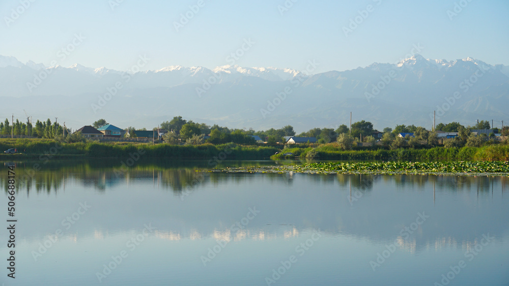 The mirror surface of the lake water reflects pink lotuses, large green water lilies, high mountains of the Trans-Ili Alatau, trees and houses. Beautiful landscape on mirror pond. Almaty, Kazakhstan