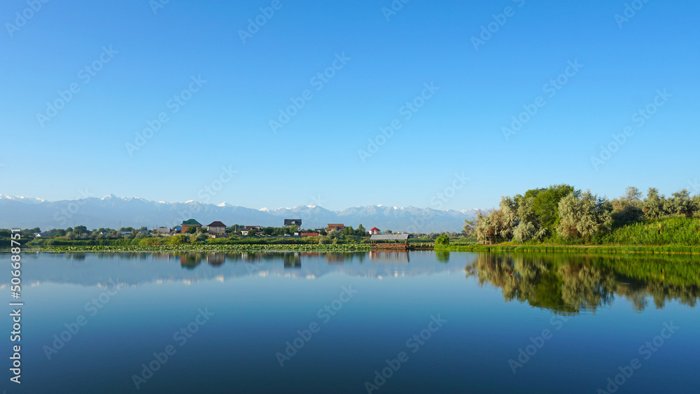 The mirror surface of the lake water reflects pink lotuses, large green water lilies, high mountains of the Trans-Ili Alatau, trees and houses. Beautiful landscape on mirror pond. Almaty, Kazakhstan