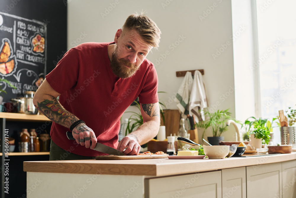Confident Caucasian man with beard on face chopping and cutting ingredients with knife on board looking at camera Confident Caucasian man with beard on face chopping and cutting ingredients with knife on board looking at camera