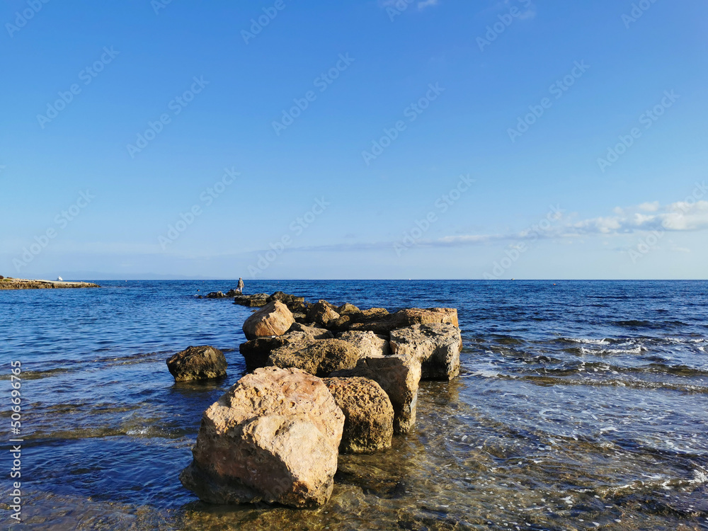 The coast of the Mediterranean Sea, a stone ridge, at the end of which is a fisherman against a blue sky with clouds.